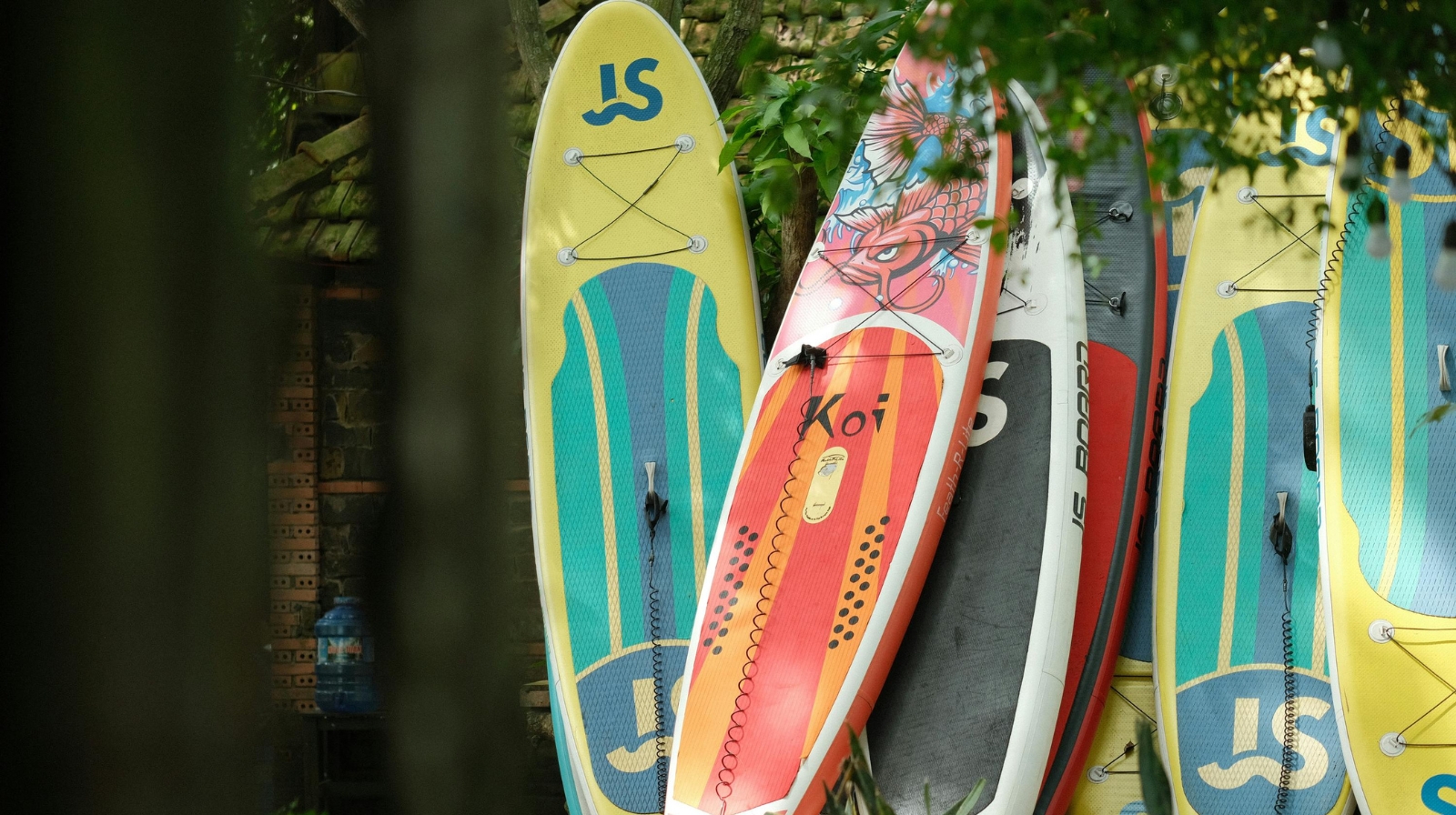 Aufgereihte Stand-Up-Paddleboards angelehnt an einer Hauswand im Grünen.