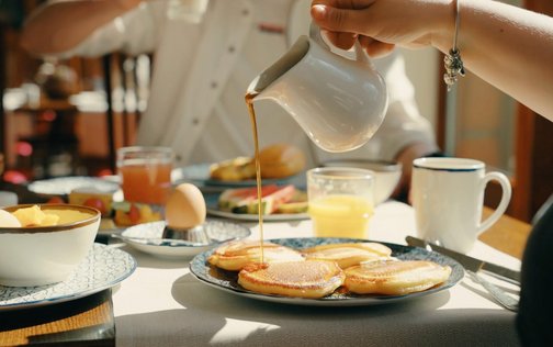 Lecker gefüllter Frühstückstisch im Sonnenschein. Brötchen, Ei, Pancakes und Ahornsirup